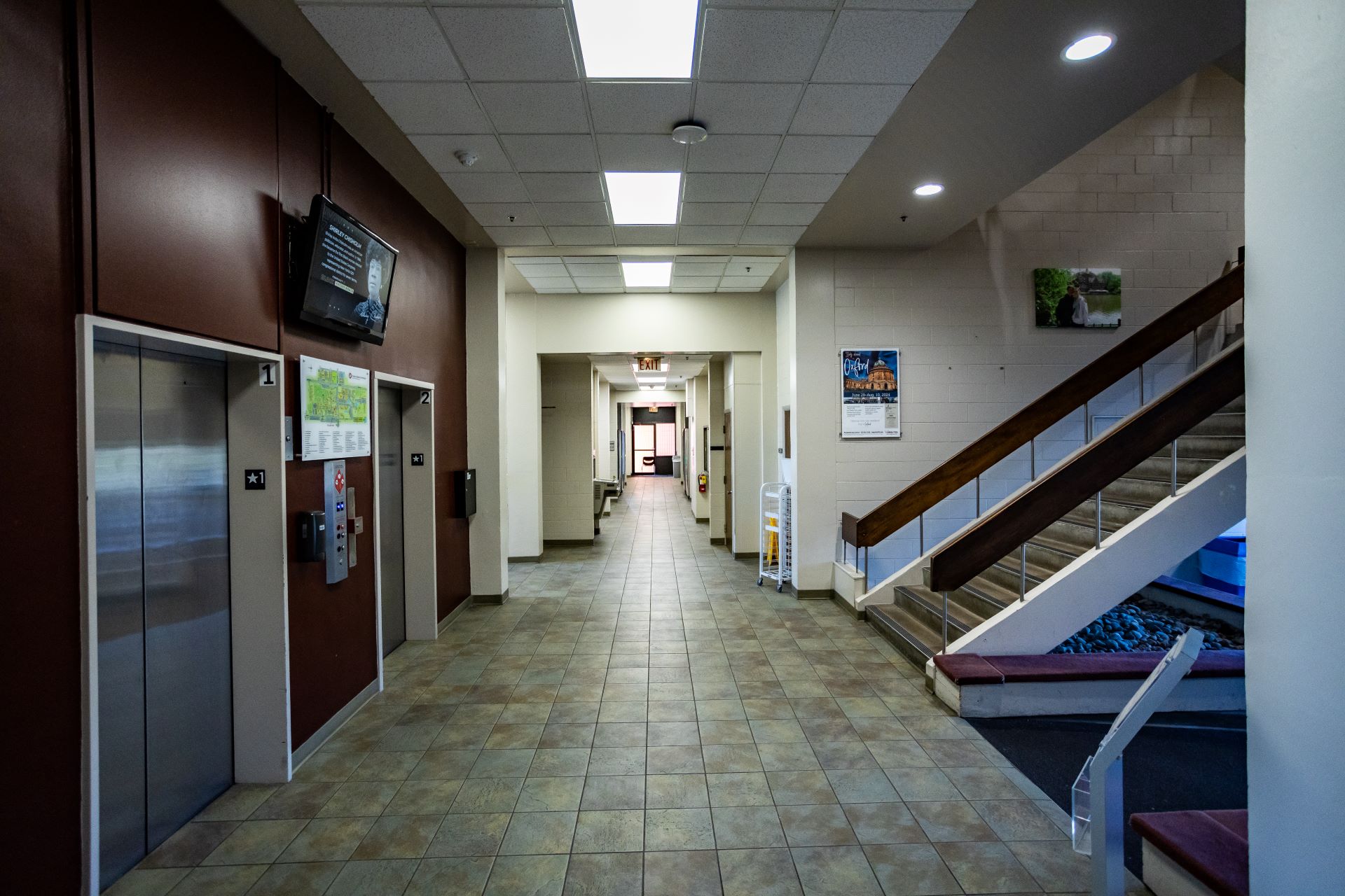 Interior hallway of a building showing two elevators on the left, a staircase on the right, and a long corridor leading to the back, with informational posters on the walls